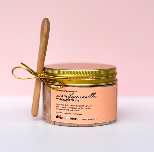 Caramélisée Vanille Macadamia sugar scrub jar with gold lid and wooden spoon tied with gold ribbon on pink backgroun