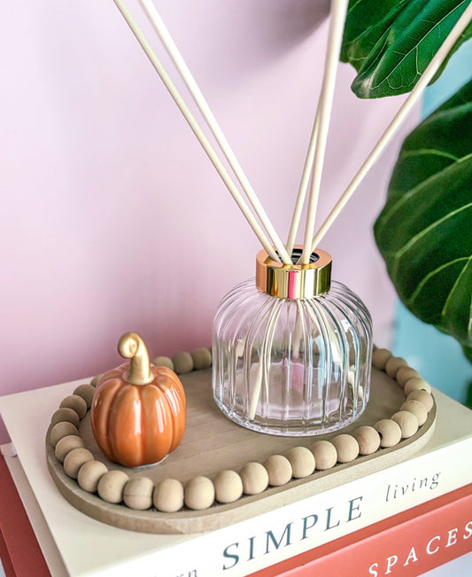 A clear ribbed glass reed diffuser with a gold collar and cream-colored reeds, styled on a stack of books next to a small orange pumpkin and a wooden bead tray.
